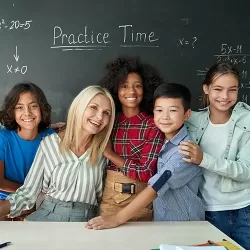 Public school teacher surrounded by students in front of chalkboard.